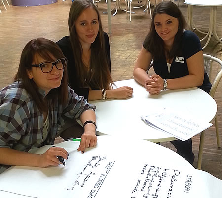 Mentoring-Workshop: Hier sind drei Mentorinnen während eines Ausbildungsworkshops zu sehen. Sie bearbeiten gerade eine Aufgabe auf Flipchart-Papier und lächeln in die Kamera